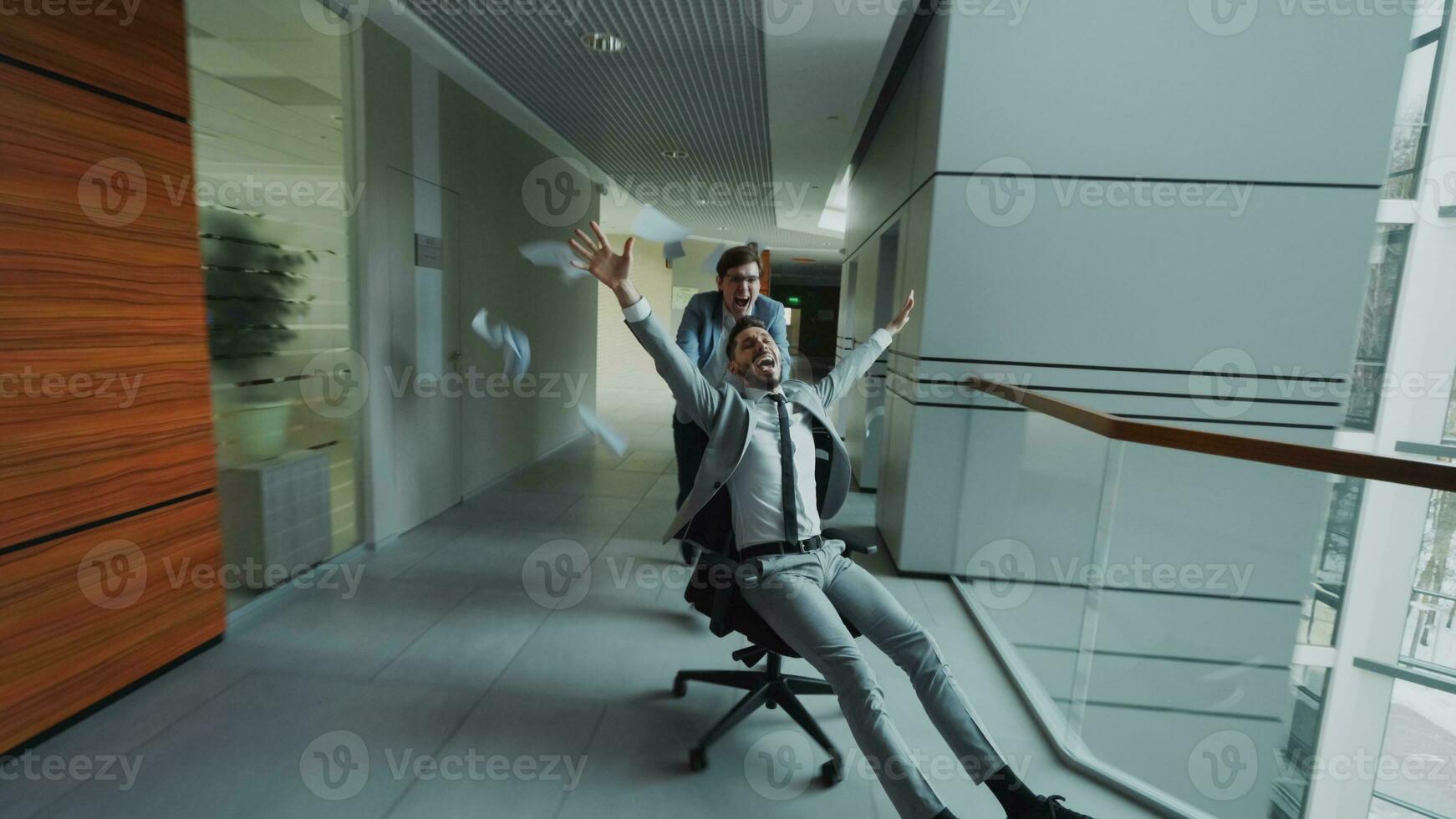 twee gek zakenlieden rijden kantoor stoel en het werpen papieren omhoog terwijl hebben pret in lobby van modern bedrijf centrum binnenshuis foto
