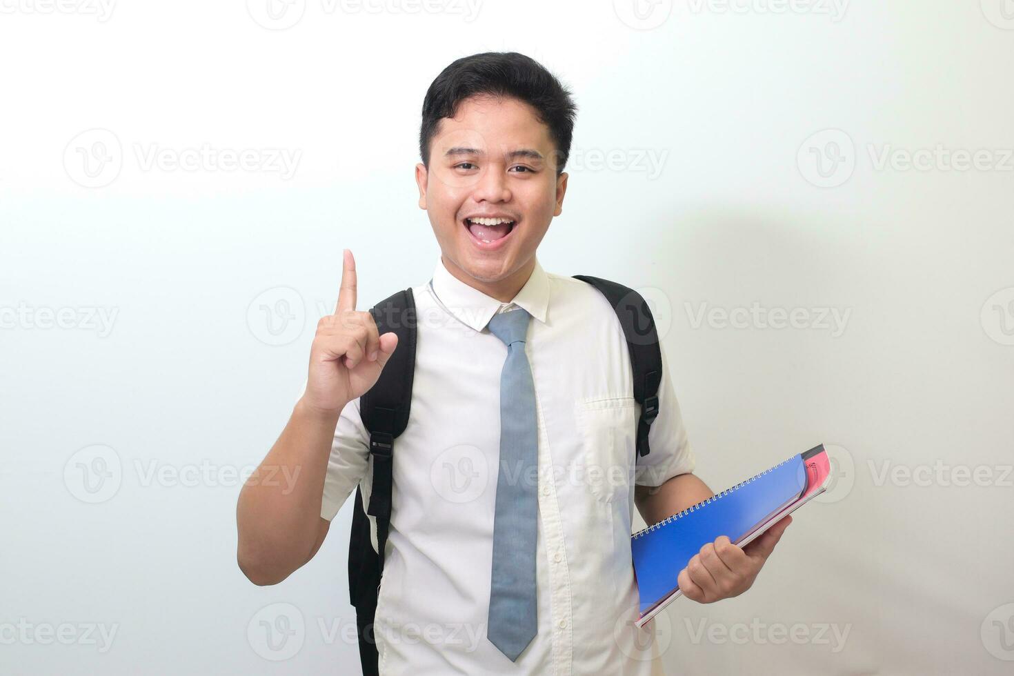 Indonesisch senior hoog school- leerling vervelend wit overhemd uniform met grijs stropdas Holding sommige boeken, glimlachen en richten omhoog met vinger herinneren iets. geïsoleerd beeld Aan wit achtergrond foto