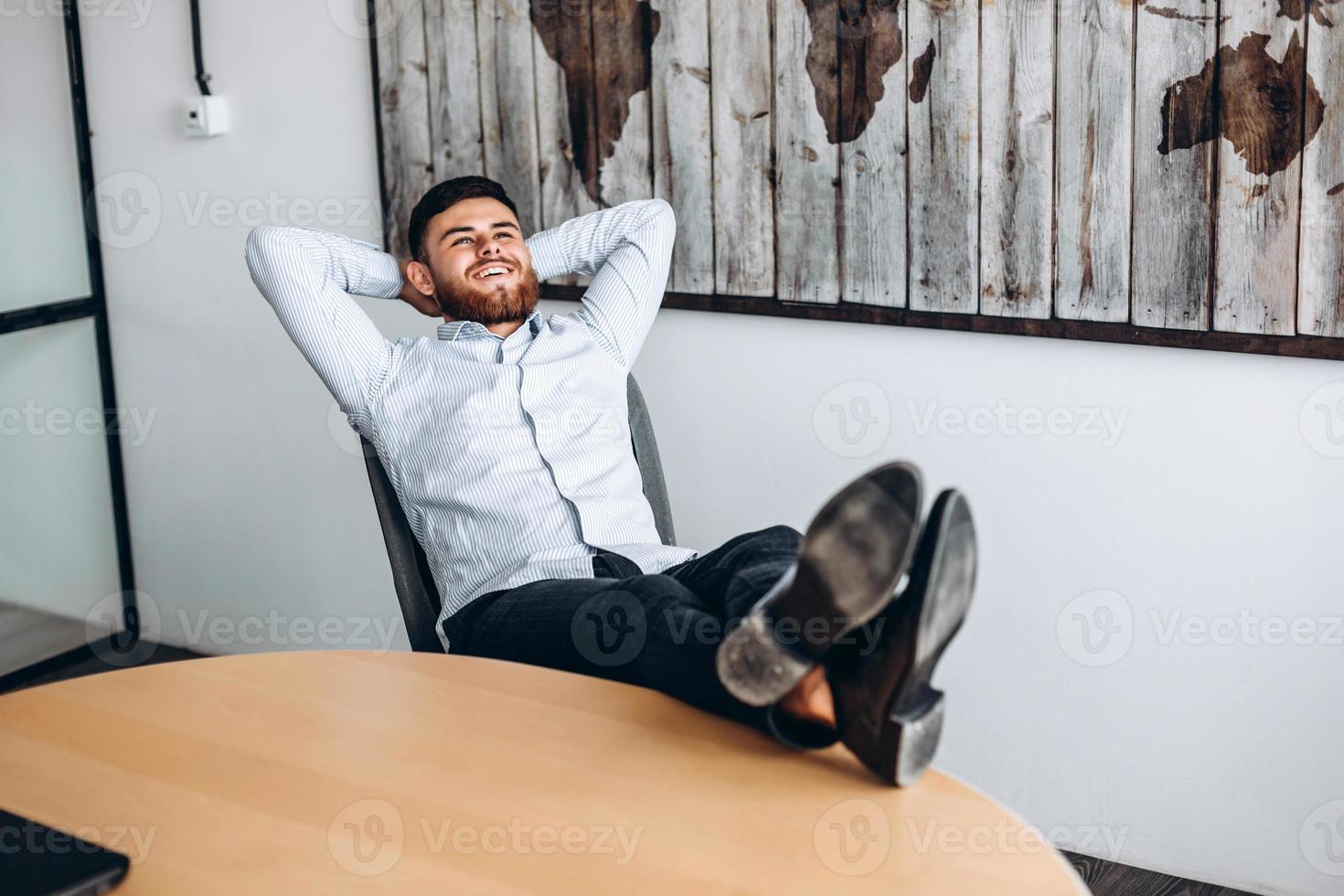 een aantrekkelijke man met een baard legt zijn handen achter zijn hoofd, zet zijn voeten op tafel en rust. foto