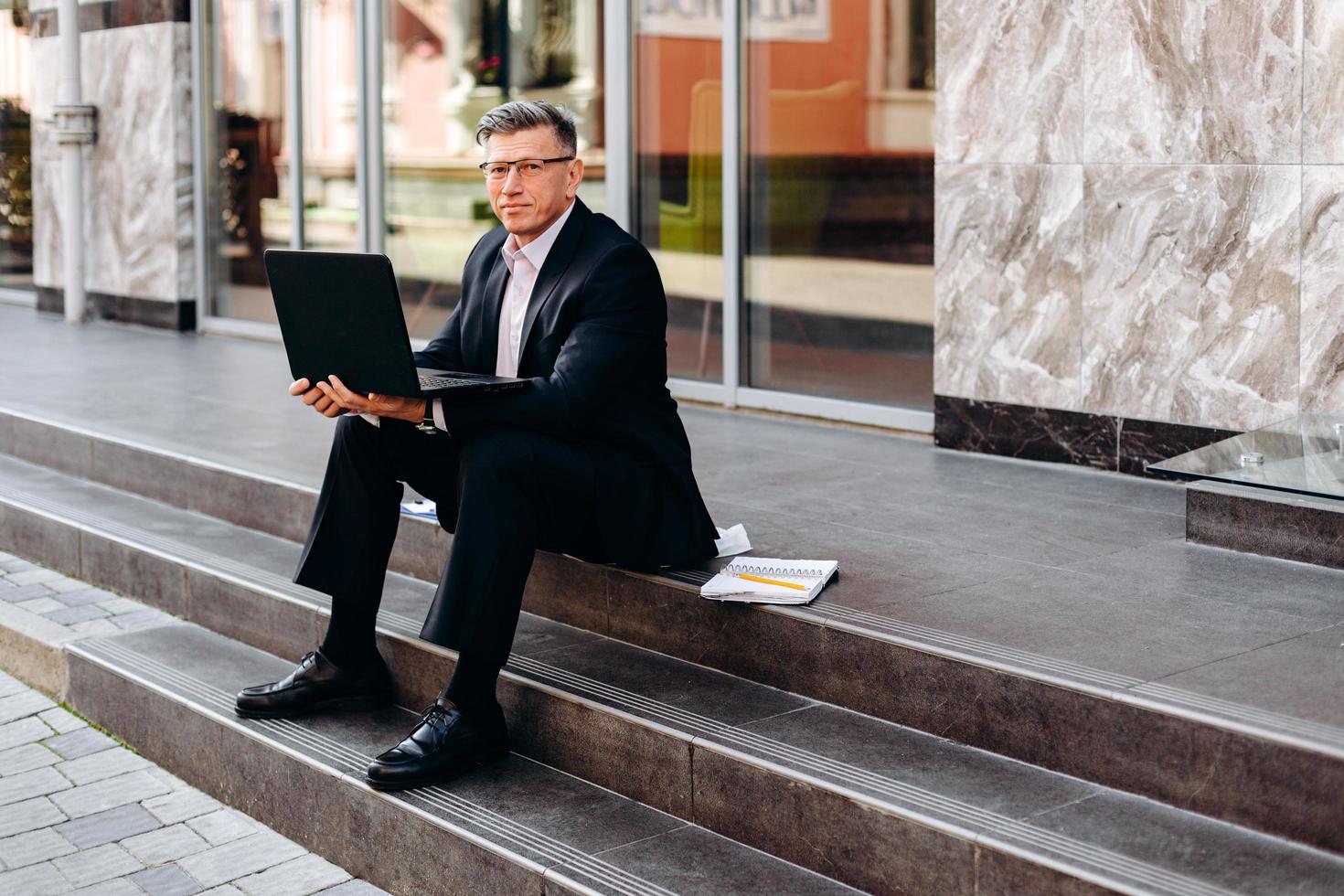 portret van senior man in pak zitten en houden van een opengeklapte laptop buiten. - afbeelding foto