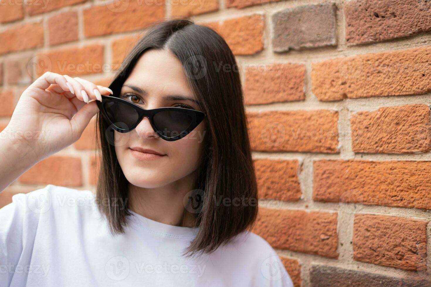 een schattig brunette meisje koket past zich aan haar zonnebril tegen een steen muur. detailopname foto