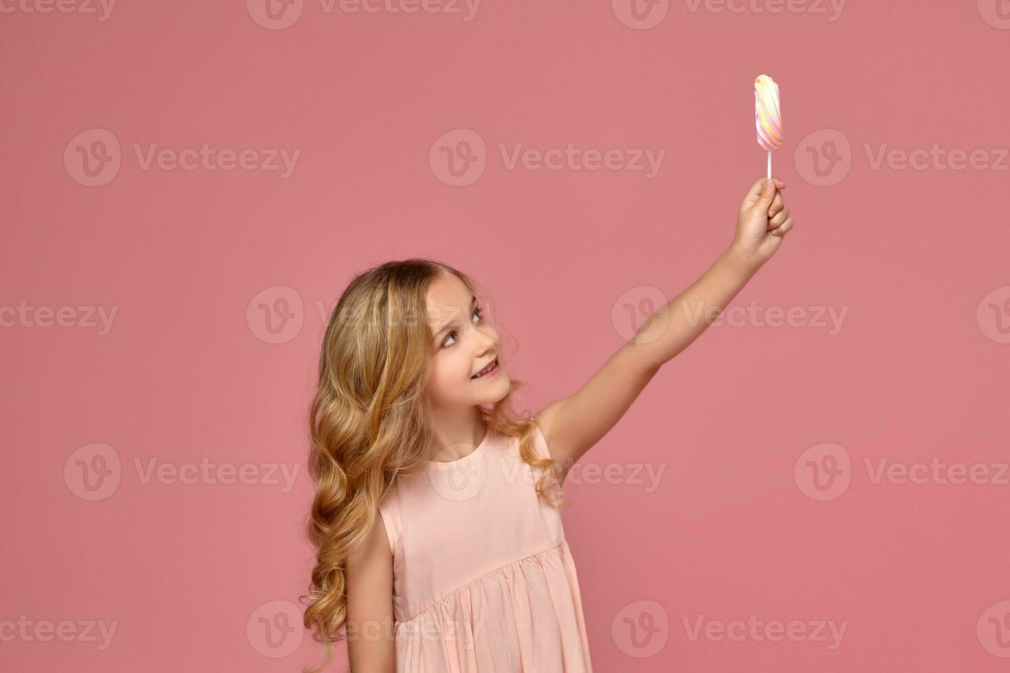 weinig meisje met een blond gekruld haar, in een roze jurk is poseren met een snoep foto