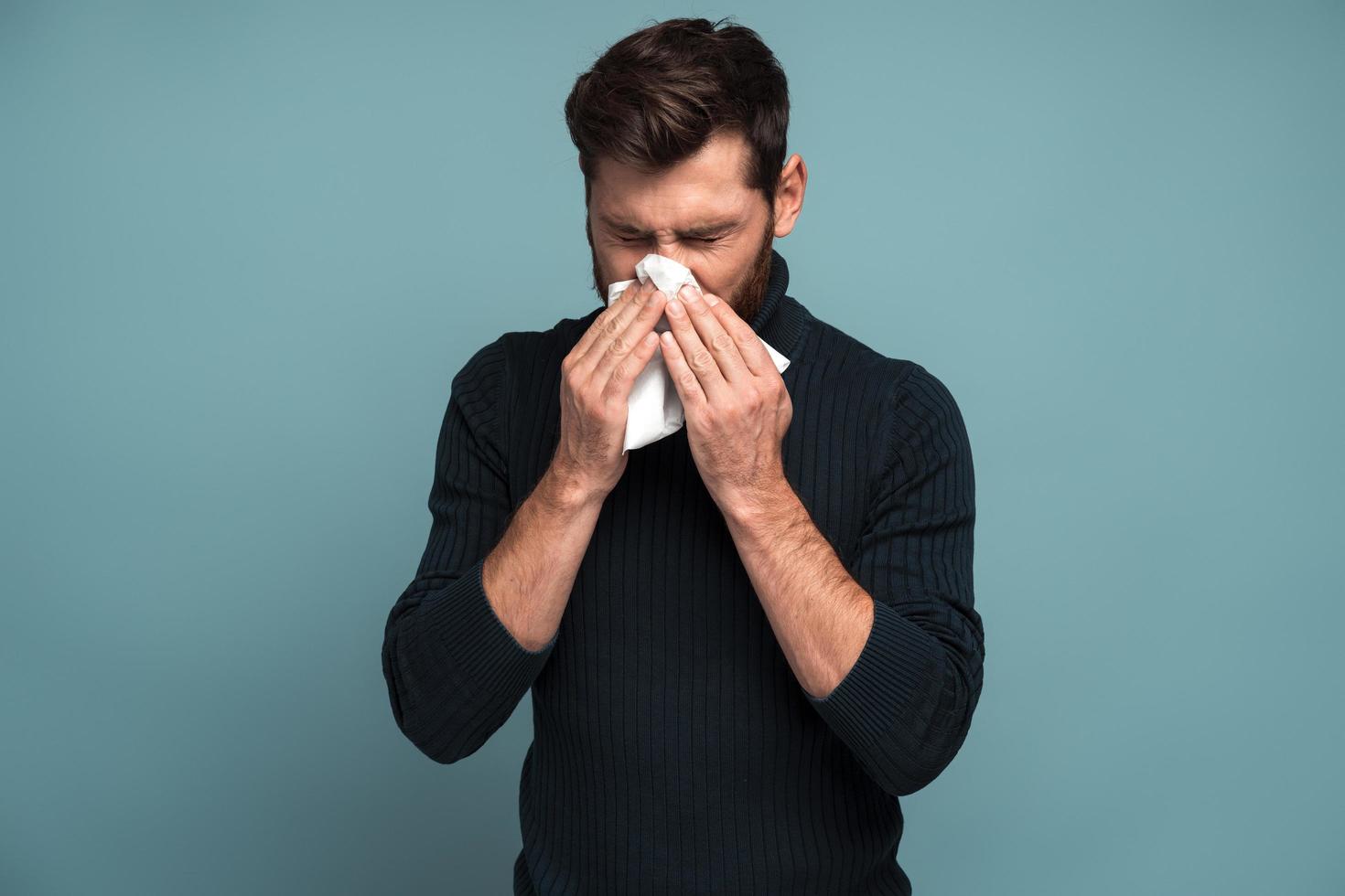 verkoudheid en griep. portret van een zieke, bebaarde jonge man die staat, weefsel op de mond houdt en verdriet en ziekte voelt. indoor studio-opname, geïsoleerd op blauwe achtergrond foto