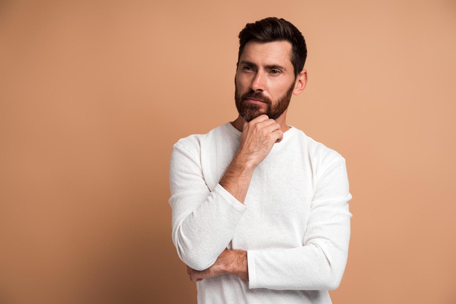 portret van een peinzende knappe bebaarde jonge man die staat, zijn gezicht aanraakt, opzij kijkt en aan iets denkt. indoor studio-opname, geïsoleerd op beige achtergrond foto