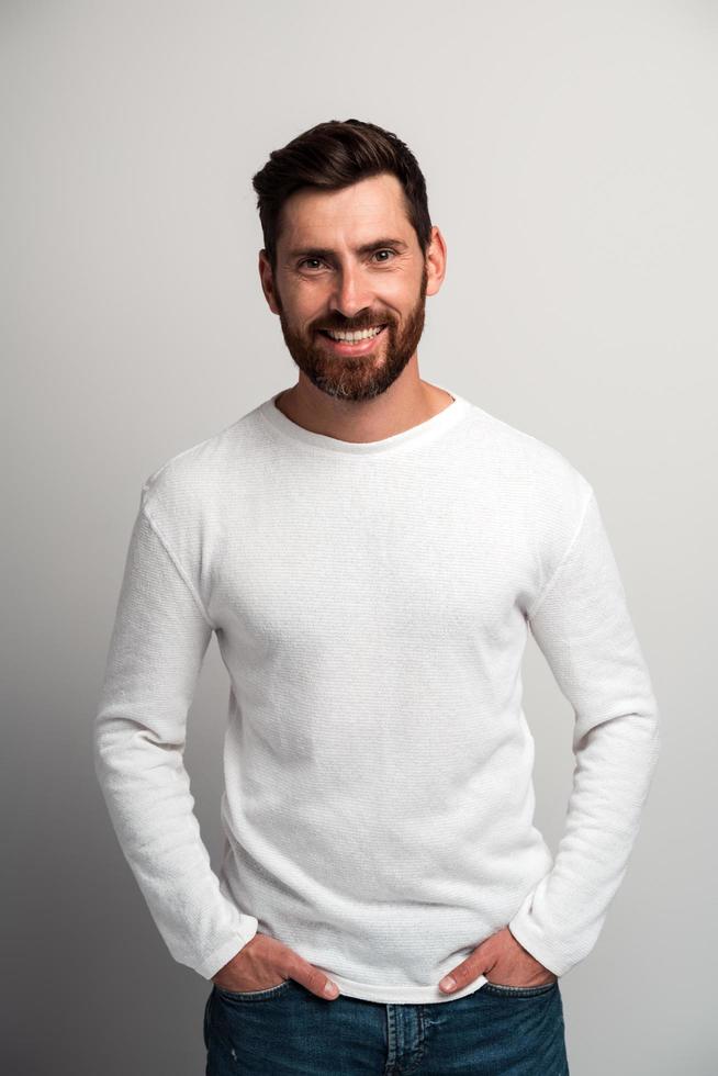 portret van een gelukkige tevreden knappe jongeman in een wit overhemd die met armen in de zakken staat en naar de camera kijkt met een brede glimlach. indoor studio-opname op witte achtergrond kopie ruimte foto