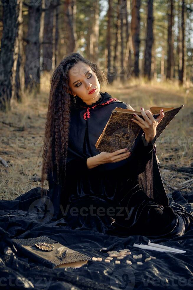 heks in zwart jurk met kaap en kap. poseren in pijnboom Woud. zittend Aan donker deken waarmee boeken, kaarsen en runen Aan het. vol lengte. foto