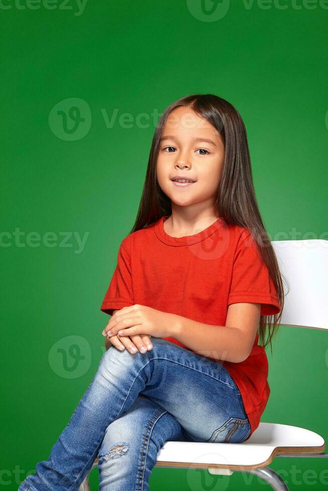 weinig meisje vervelend rood t-kort en poseren Aan stoel foto