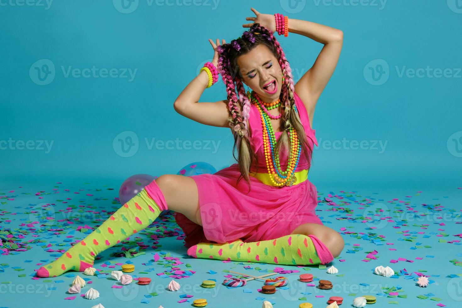 lief meisje met een veelkleurig vlechtjes kapsel en helder verzinnen, poseren in studio met lucht ballonnen en confetti tegen een blauw achtergrond. foto