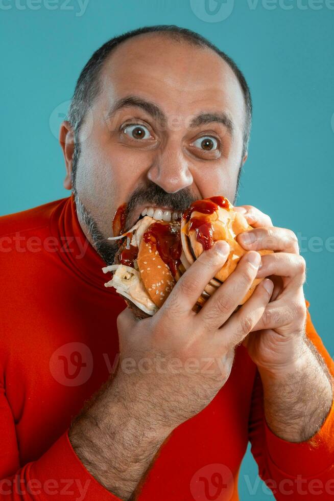 detailopname portret van een middelbare leeftijd Mens met baard, gekleed in een rood coltrui, poseren met hamburgers tegen een blauw achtergrond. snel voedsel. foto