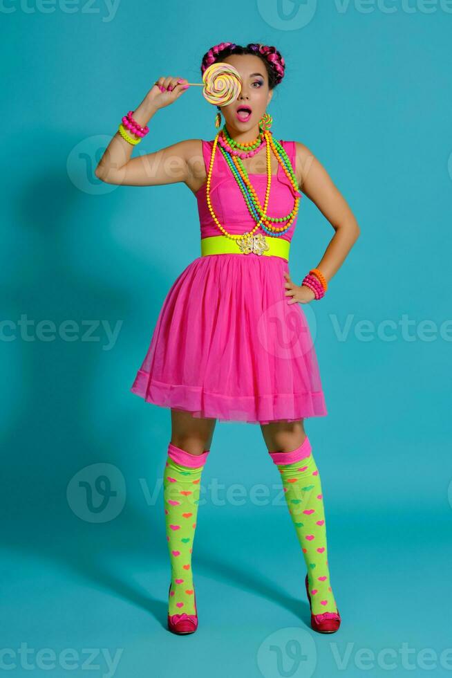 lief meisje met een veelkleurig vlechtjes kapsel en helder verzinnen, poseren in studio tegen een blauw achtergrond, Holding een lolly in haar hand. foto