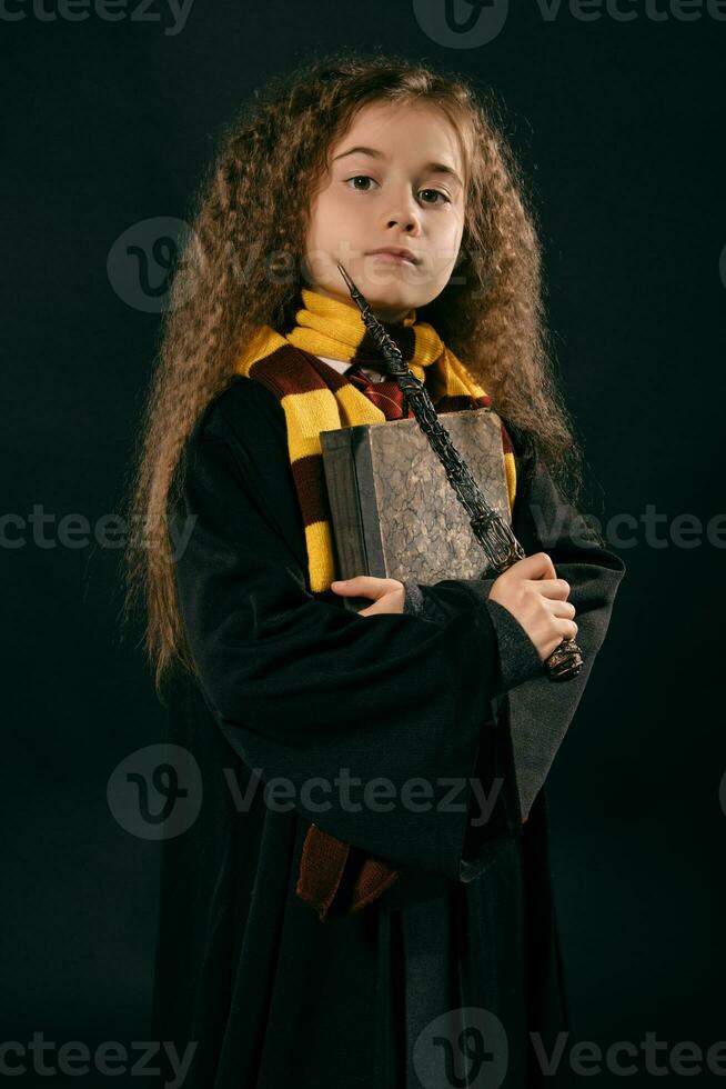 portret van een weinig heks meisje met lang bruin haar- gekleed in donker jas, Holding magie toverstaf in haar hand, poseren Aan zwart studio achtergrond. foto