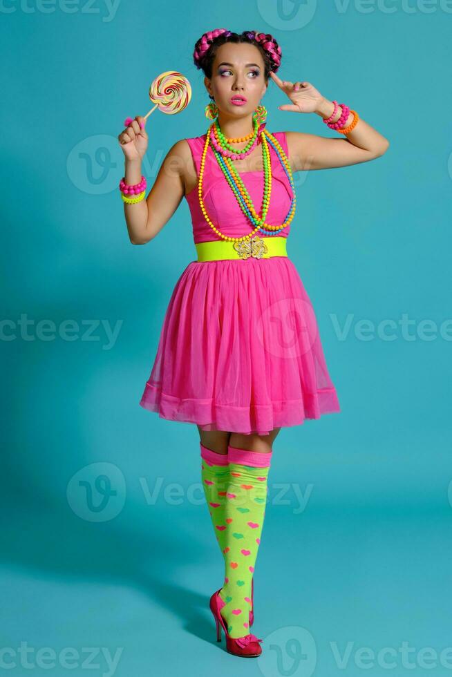 lief meisje met een veelkleurig vlechtjes kapsel en helder verzinnen, poseren in studio tegen een blauw achtergrond, Holding een lolly in haar hand. foto