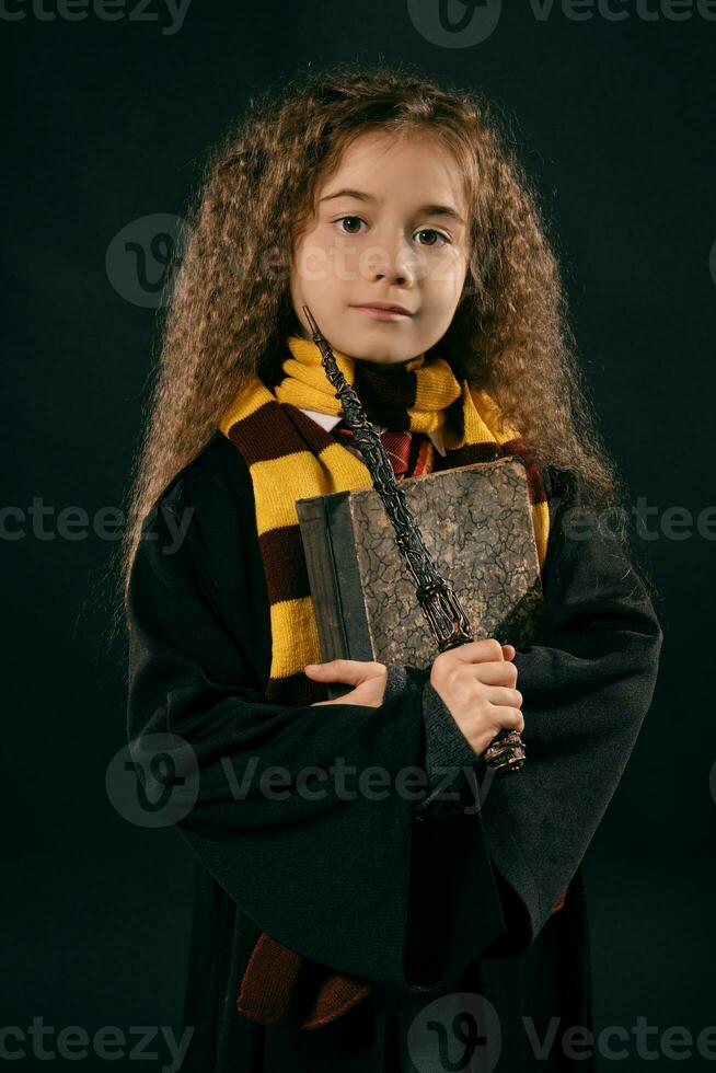 portret van een weinig heks meisje met lang bruin haar- gekleed in donker jas, Holding magie toverstaf in haar hand, poseren Aan zwart studio achtergrond. foto