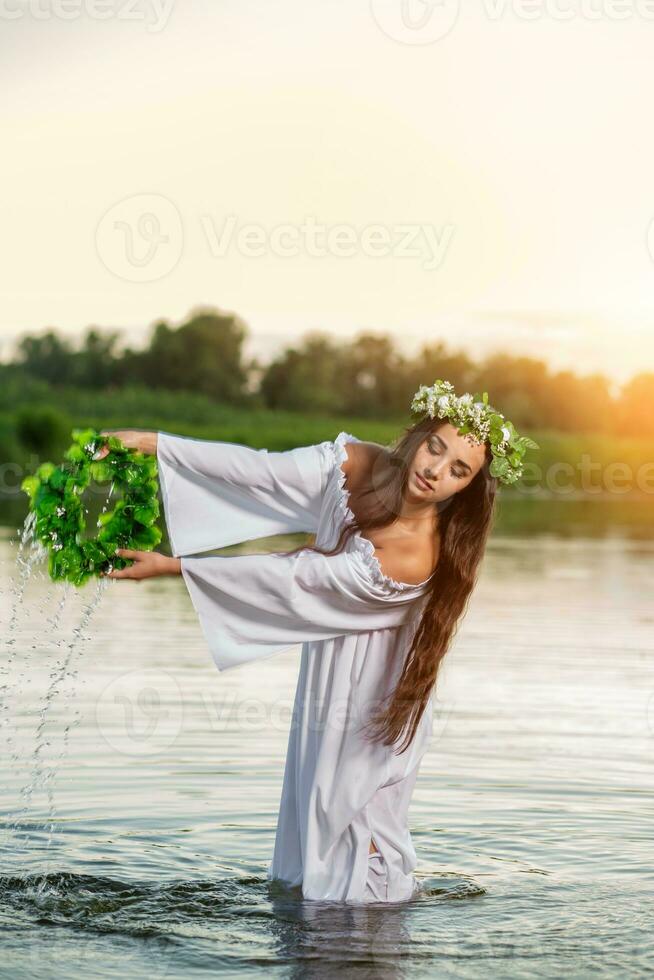 mooi zwart haren meisje in wit wijnoogst jurk en krans van bloemen staand in water van meer. zon gloed. foto