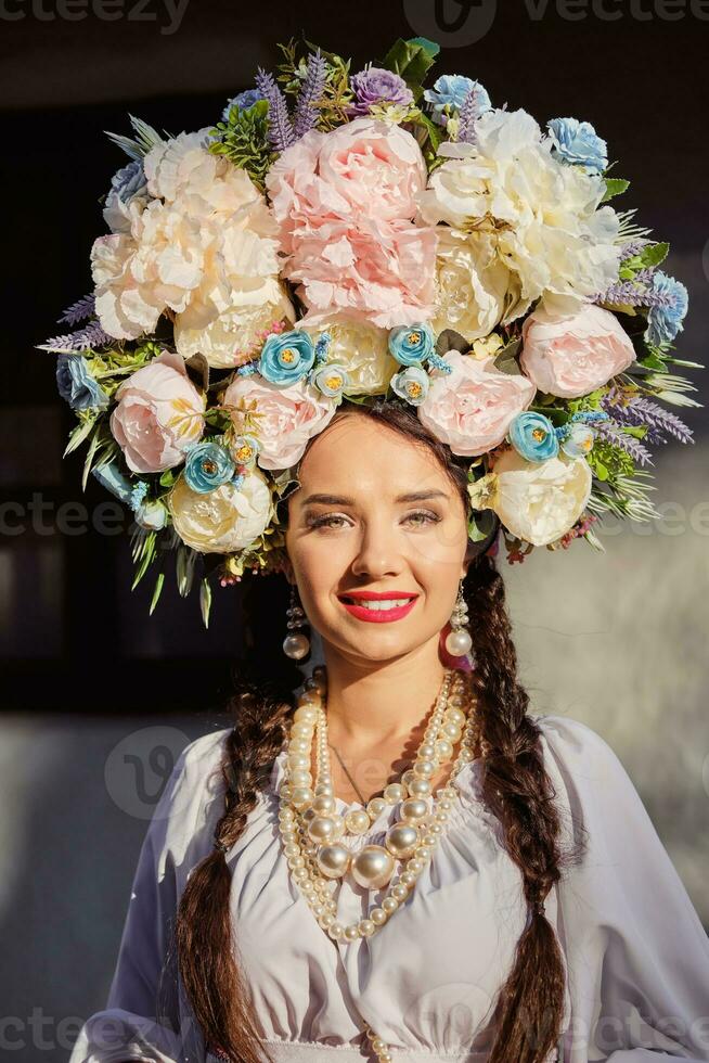 brunette meisje in een wit oekraïens authentiek nationaal kostuum en een krans van bloemen is poseren tegen een wit hut. detailopname. foto