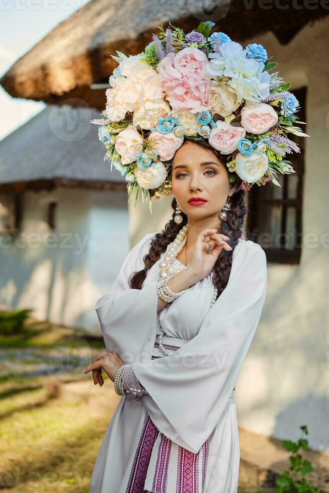 brunette meisje in een wit oekraïens authentiek nationaal kostuum en een krans van bloemen is poseren tegen een wit hut. detailopname. foto