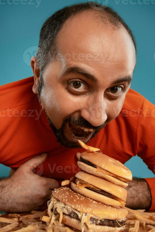 detailopname portret van een middelbare leeftijd Mens met baard, gekleed in een rood coltrui, poseren met hamburgers en Frans Patat. blauw achtergrond. snel voedsel. foto