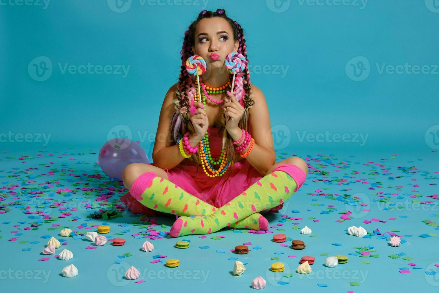 lief meisje met een veelkleurig vlechtjes kapsel en helder verzinnen, poseren in studio met lolly, lucht ballonnen en confetti tegen een blauw achtergrond. foto