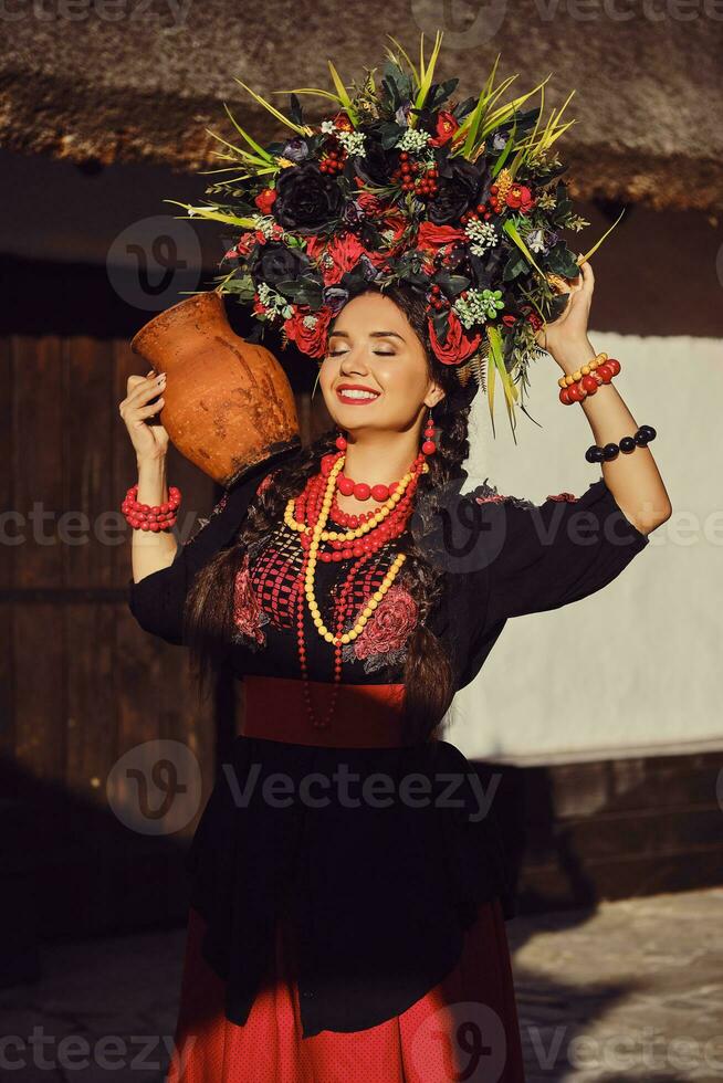 brunette meisje in een zwart en rood oekraïens geborduurd authentiek nationaal kostuum en een krans van bloemen is poseren staand Bij de poort. foto