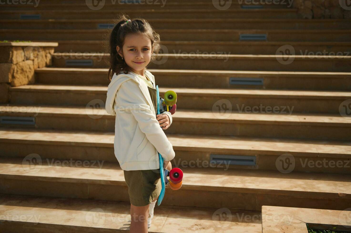 Kaukasisch gelukkig glimlachen elementair leeftijd school- kind schaatser met skateboard buitenshuis. actief weinig meisje met cent bord foto