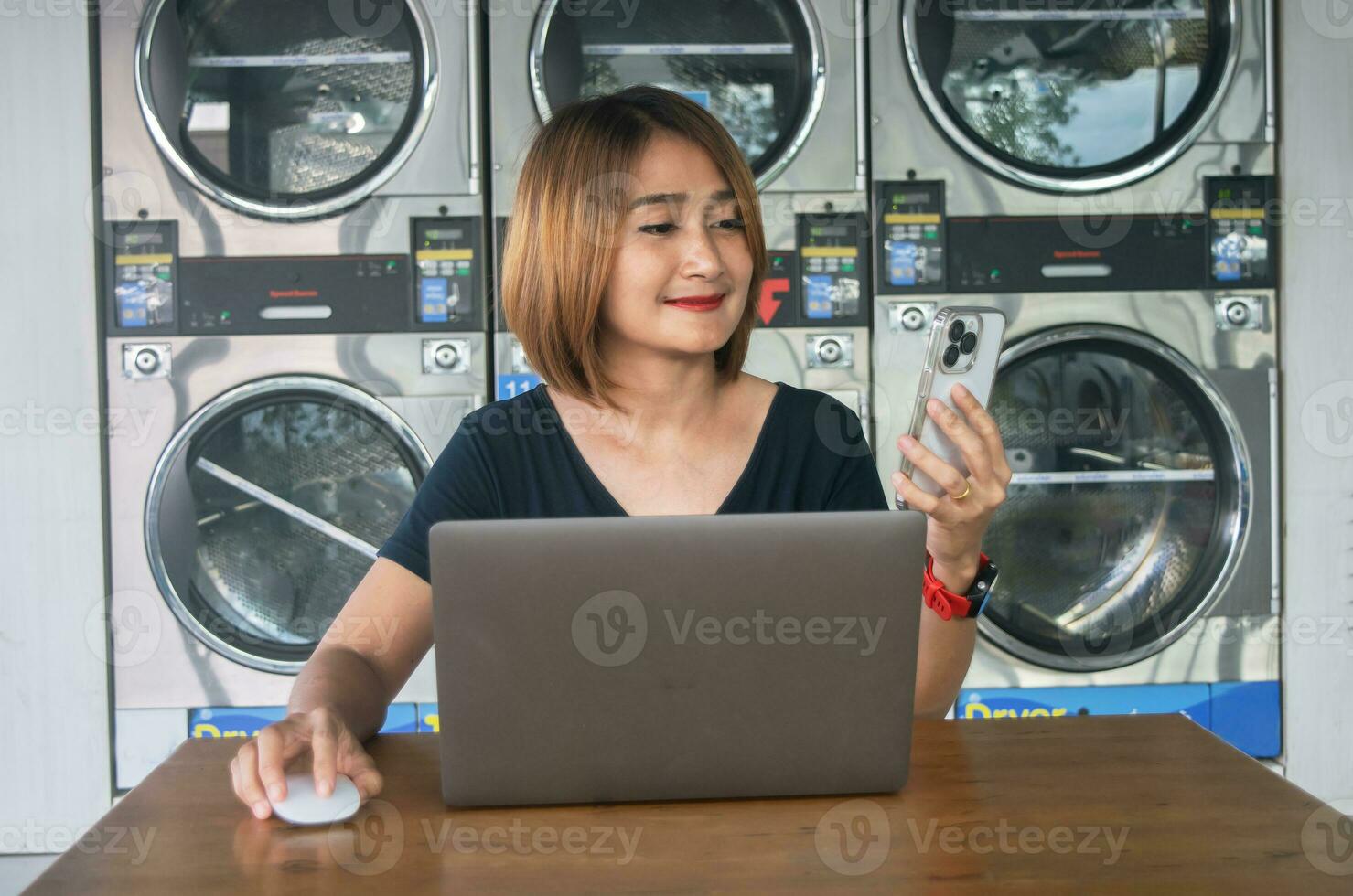Dames Holding smartphone video telefoontje met vriend en gebruik makend van laptop naar werk in de wasserij Oppervlakte terwijl aan het wachten voor het wassen machine foto
