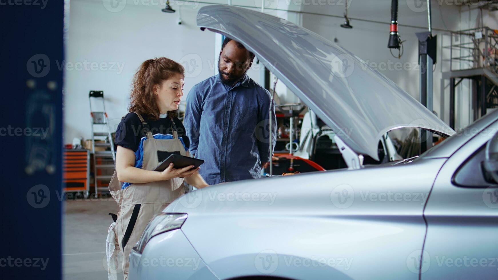 gecertificeerd deskundige in garage gebruik makend van laptop naar volgen checklist terwijl aan het doen onderhoud Aan voertuig, pratend met cliënt. monteur in auto onderhoud doet controle Aan auto, dolly in schot foto