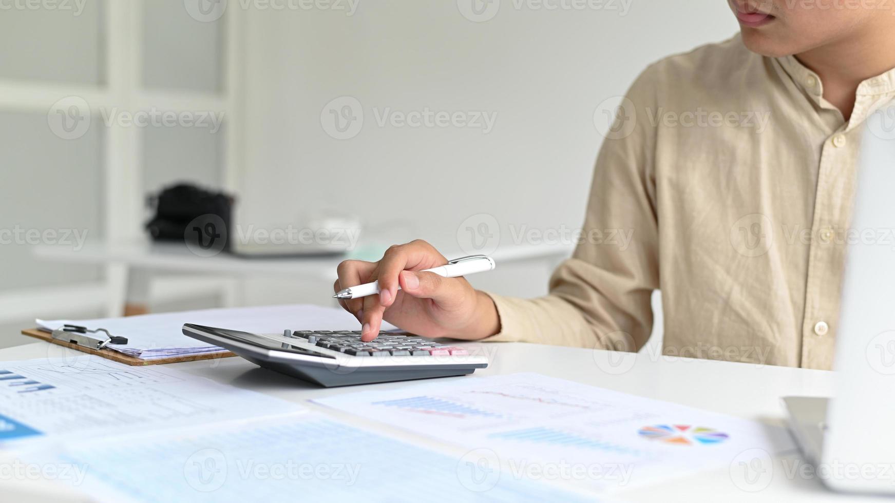 een jonge man met een pen in de hand bedient een rekenmachine, grafische gegevens en laptop op het bureau. foto