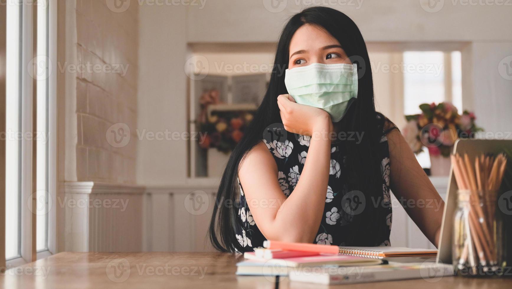 vermoeide vrouw met een medisch masker kijkt uit het raam, met laptop en briefpapier op tafel. foto