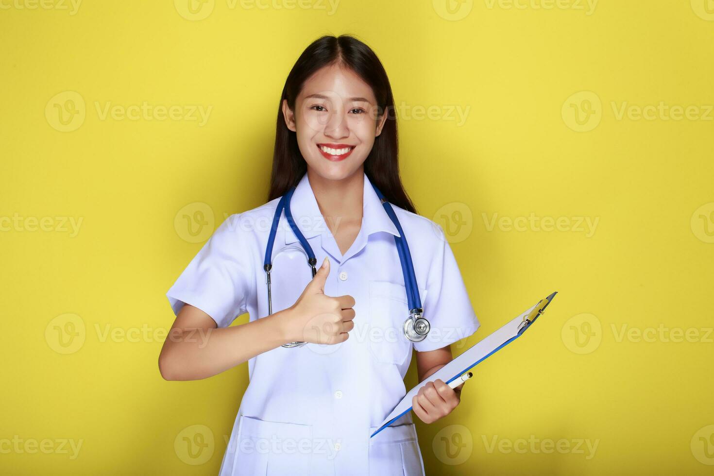 een jong vrouw vervelend een dokter uniform poses met een duimen omhoog en blijft hangen een medisch stethoscoop, portret van een mooi jong vrouw in een geel achtergrond, foto