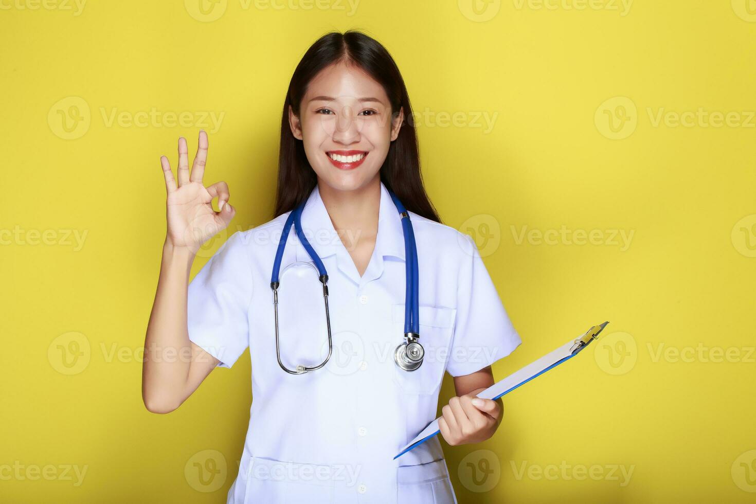 portret van een mooi jong vrouw in een geel achtergrond, mooi Aziatisch vrouw staand gelukkig maken OK gebaar met handen en vervelend dokter uniform. foto