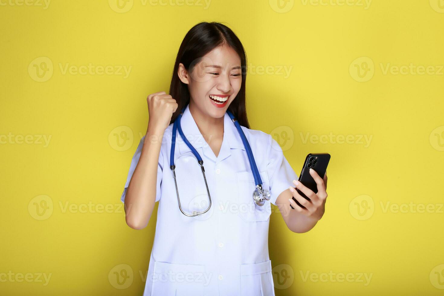 portret van een mooi jong vrouw in een geel achtergrond, Aziatisch vrouw poses met een cel telefoon terwijl vervelend een dokter uniform en een stethoscoop. foto