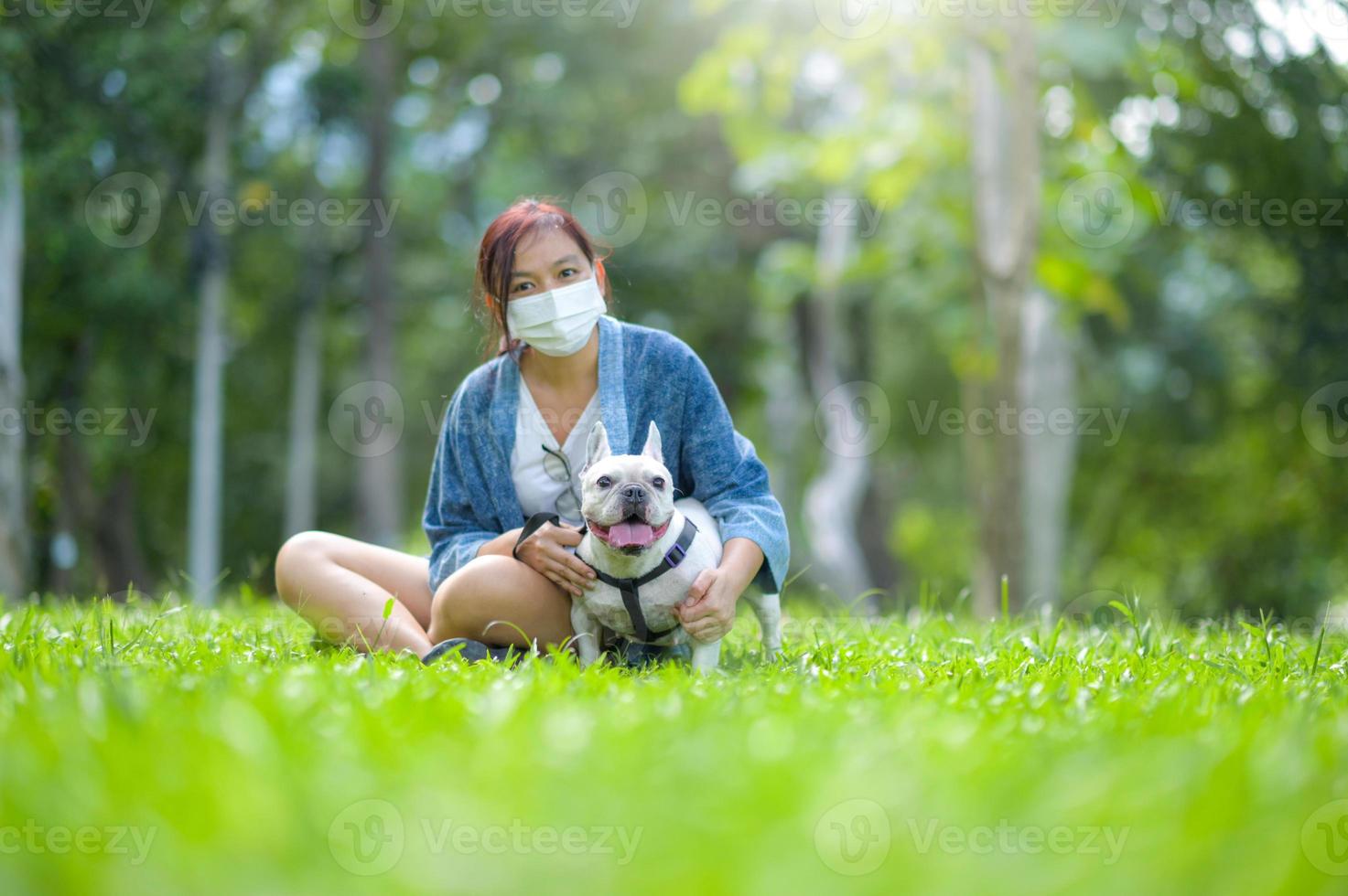 een vrouw met een medisch masker zit op het gazon met een witte franse bulldog. foto