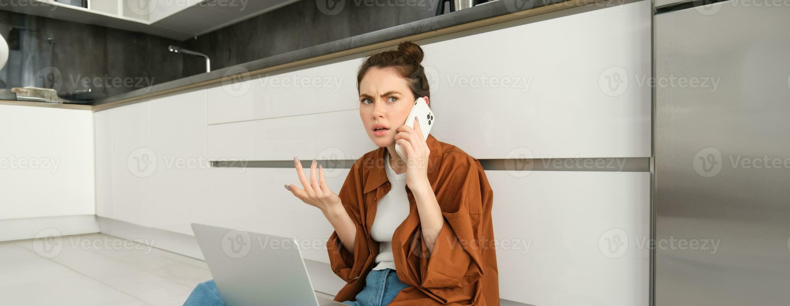 vrouw met gefrustreerd en verward gezicht pratend Aan mobiel telefoon, schouderophalend en fronsend, horen slecht nieuws over- de telefoon, zit Aan verdieping met laptop, oproepen klant ondersteuning foto