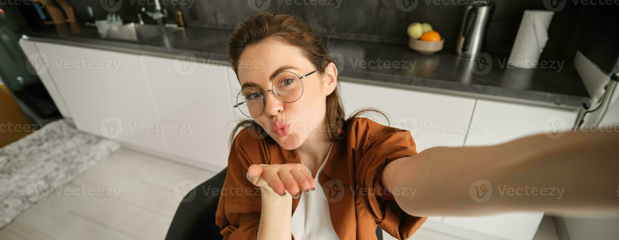 portret van elegant jong vrouw Bij huis, nemen selfie Aan smartphone, poseren in keuken en glimlachen Bij mobiel camera foto