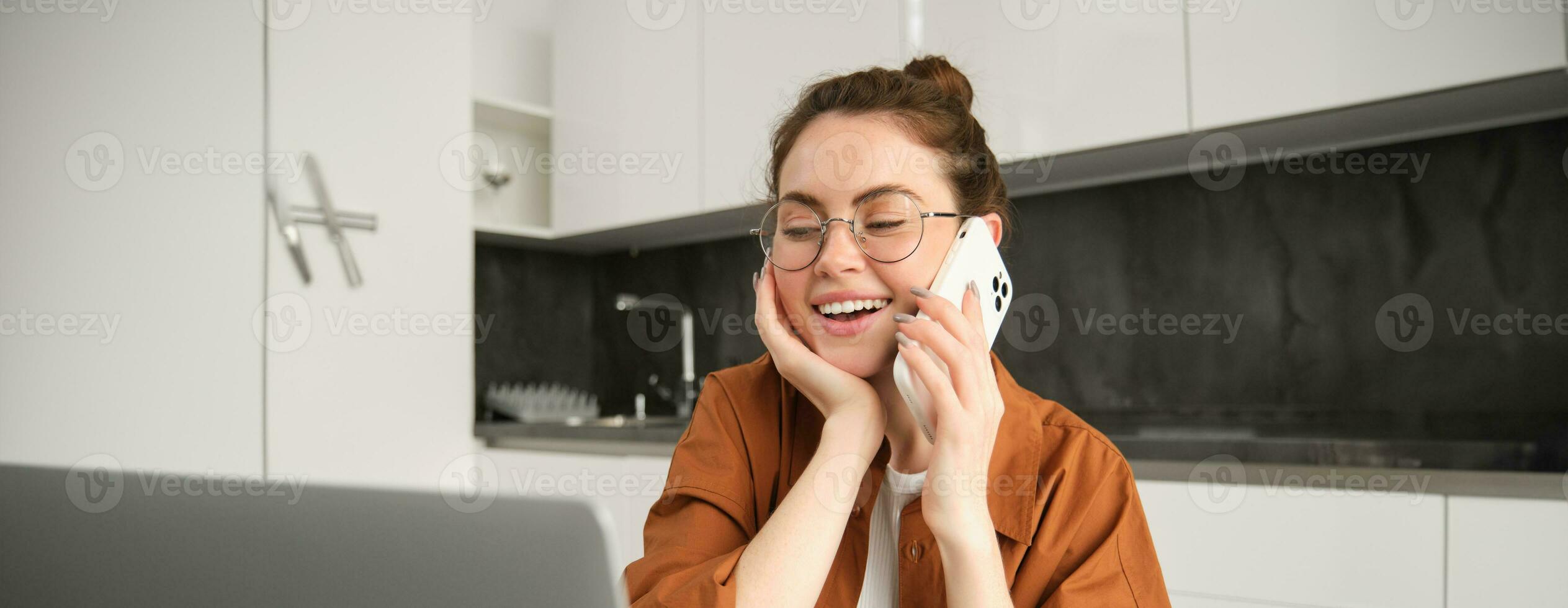 portret van mooi jong vrouw beheren haar eigen bedrijf van huis, freelancer maken telefoon telefoongesprek, zittend in keuken met laptop en glimlachen foto