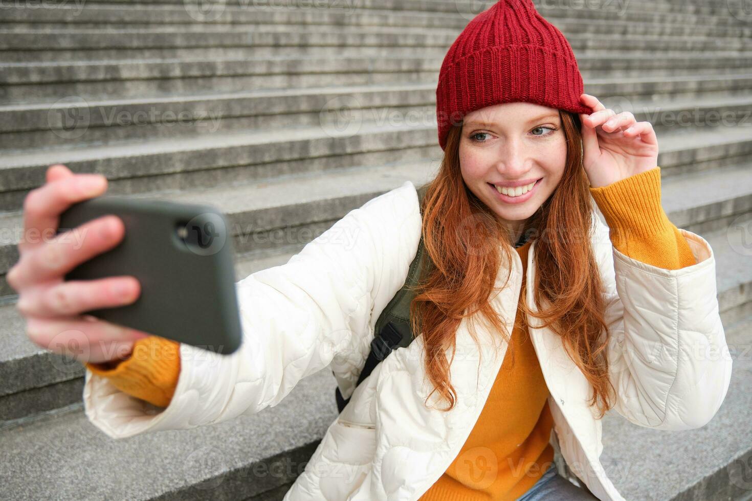 elegant jong meisje in rood hoed, duurt foto's Aan smartphone camera, maakt selfie net zo ze zit Aan trap in de buurt museum, poseren voor foto met app filter