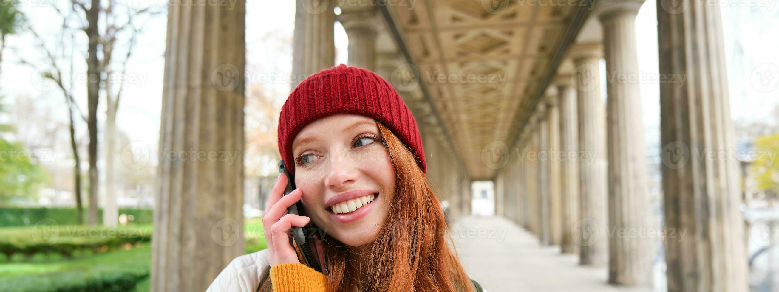 glimlachen schattig roodharige vrouw maakt een telefoon telefoongesprek, houdt telefoon in de buurt jaar, heeft mobiel gesprek, gebruik makend van smartphone Aan straat foto