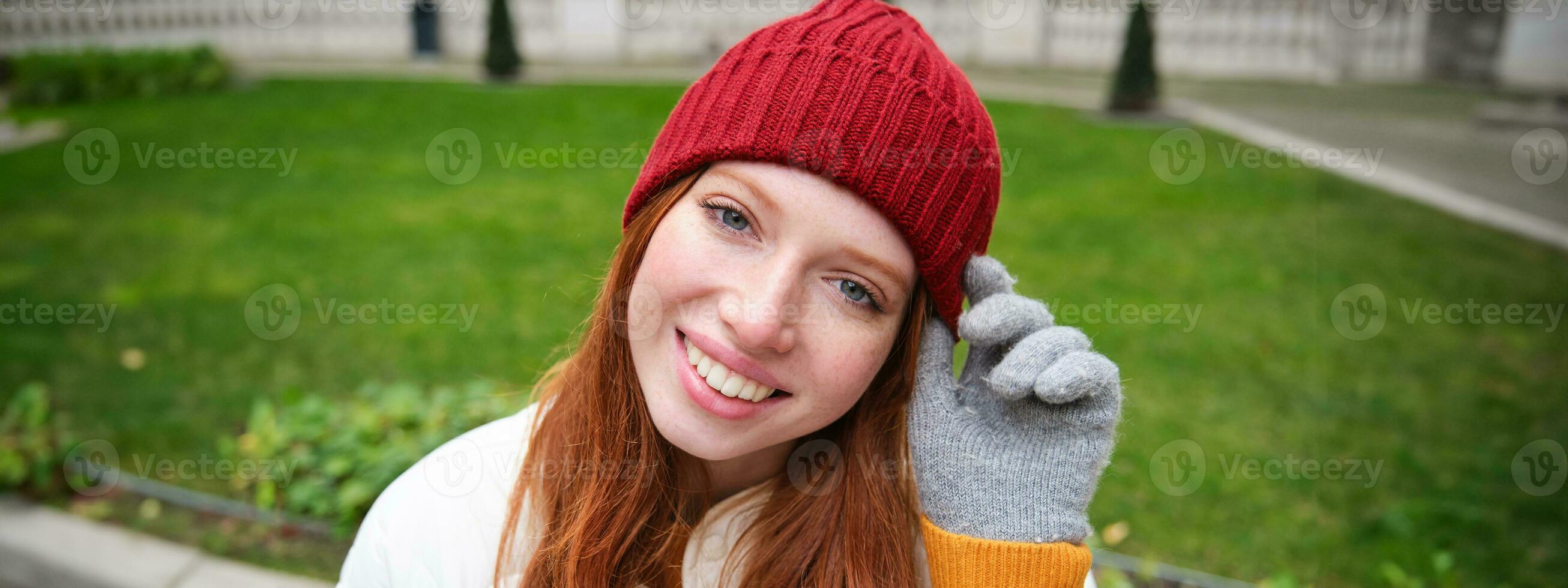 dichtbij omhoog portret van mooi roodharige vrouw in rood gebreid hoed, warm handschoenen, glimlachen en op zoek gelukkig Bij camera, zittend in park foto