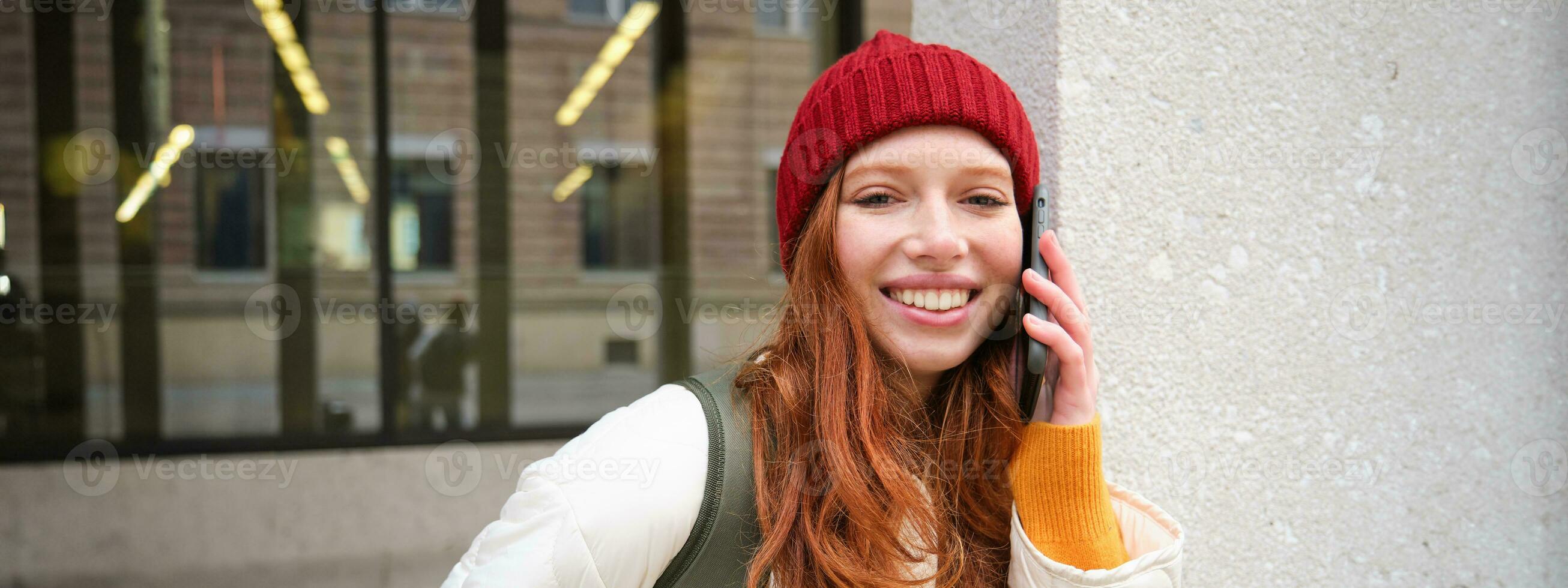 jong mensen en mobiel verbinding. gelukkig roodharige meisje praat Aan telefoon, maakt telefoon telefoongesprek, staat buitenshuis met rugzak en toepassingen smartphone app foto