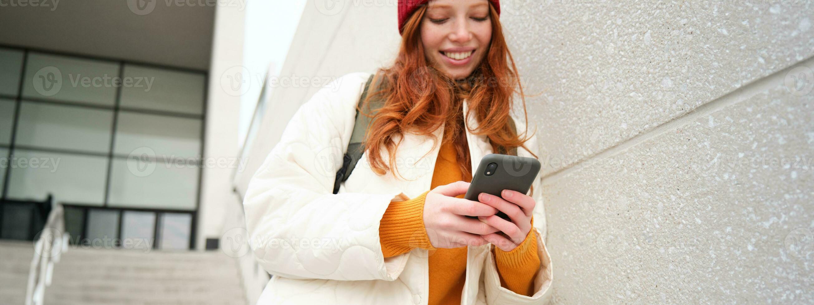roodharige meisje, jong vrouw toerist met rugzak, houdt smartphone, looks voor route Aan mobiel sollicitatie, zoekopdrachten voor hotel Aan telefoon kaart, glimlacht gelukkig foto