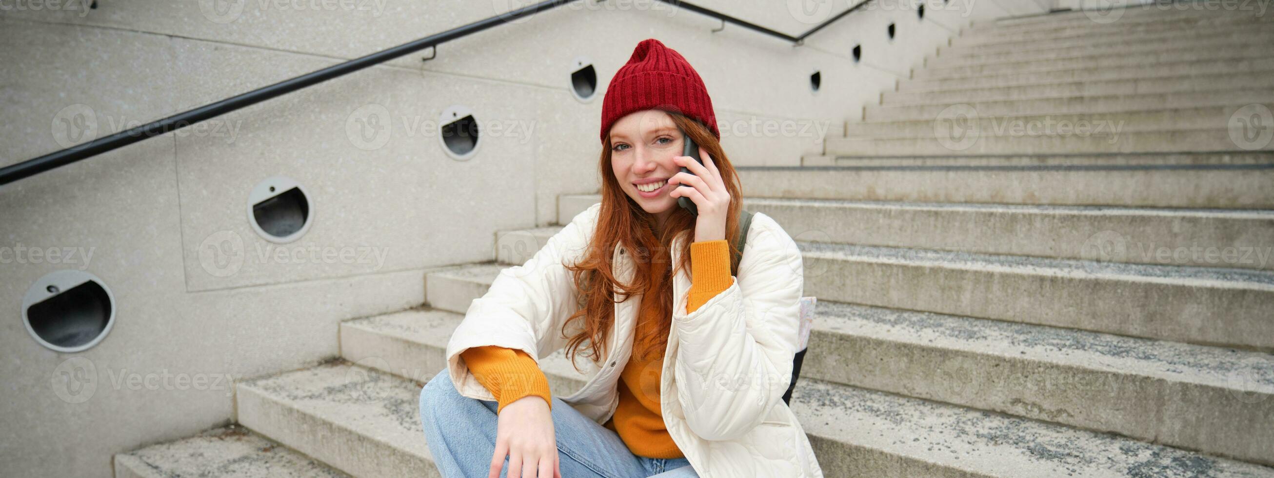 jong elegant roodharige meisje in rood hoed, zit Aan straat en praat Aan mobiel telefoon, heeft telefoon gesprek, ringen haar vriend terwijl ontspant buitenshuis foto