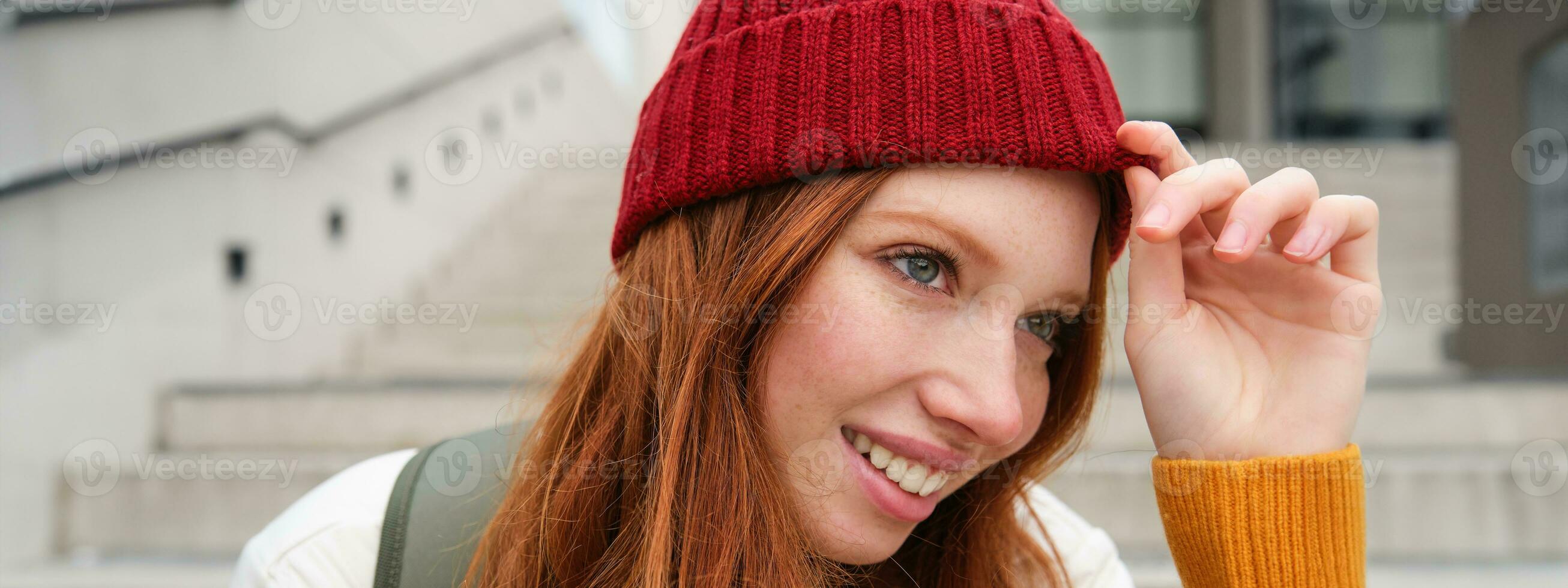 elegant roodharige meisje in warm rood hoed, glimlachen ontspannen, zittend met rugzak Aan trap in de buurt gebouw, wacht voor iemand buitenshuis foto