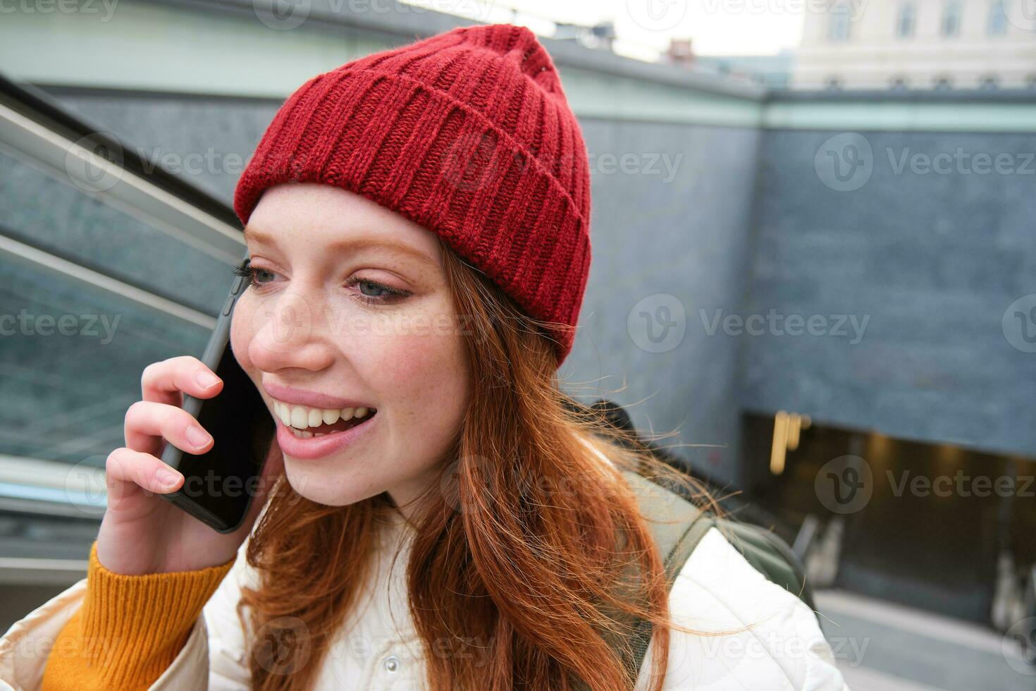 portret van jong roodharige vrouw wandelingen in de omgeving van stad, gaat omhoog trap met mobiel telefoon, praat Aan smartphone en glimlacht foto