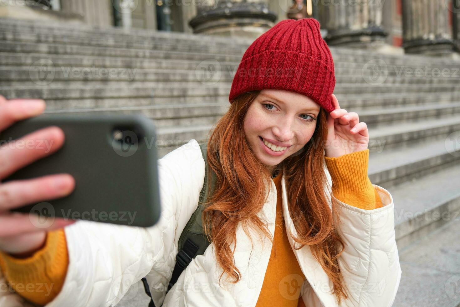 elegant jong meisje in rood hoed, duurt foto's Aan smartphone camera, maakt selfie net zo ze zit Aan trap in de buurt museum, poseren voor foto met app filter