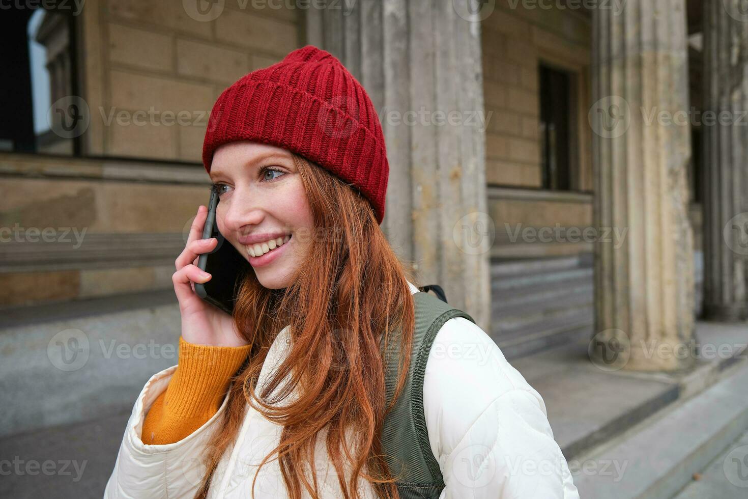 glimlachen jong roodharige vrouw luistert naar stem bericht, maakt een telefoon telefoongesprek, wandelingen Aan straat en praat naar iemand Aan smartphone foto