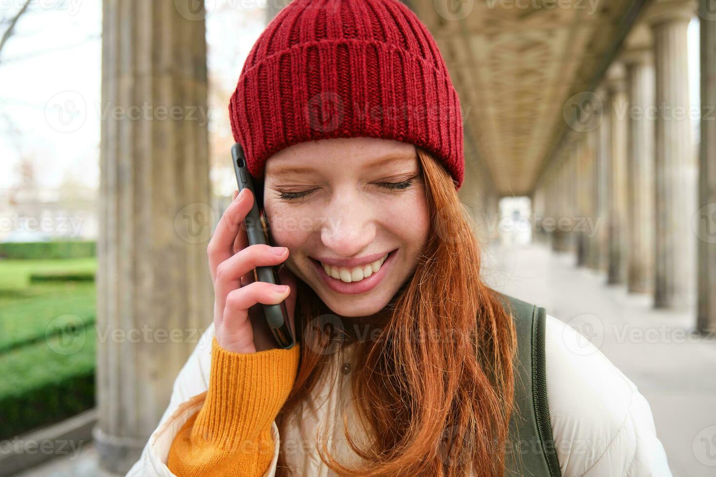 portret van roodharige Europese meisje in rood hoed, maakt een telefoon telefoongesprek, wandelingen in stad en praat naar vriend Aan smartphone foto