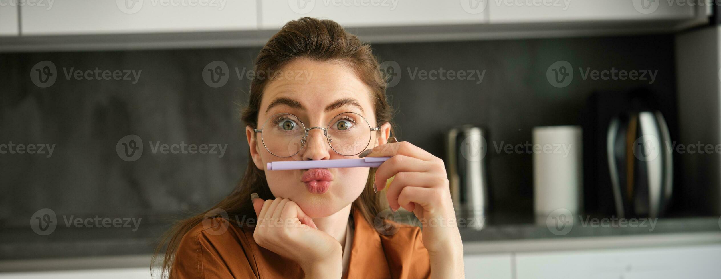 dichtbij omhoog portret van grappig, dwaas jong vrouw, spelen met pen, Holding potlood Aan top van haar lip en grimassen, zittend in keuken foto