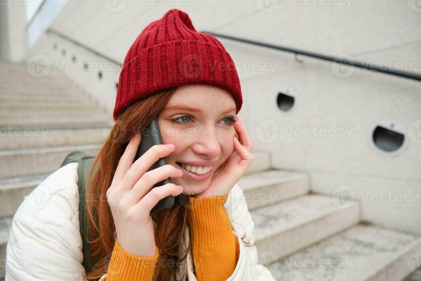 jong elegant roodharige meisje in rood hoed, zit Aan straat en praat Aan mobiel telefoon, heeft telefoon gesprek, ringen haar vriend terwijl ontspant buitenshuis foto