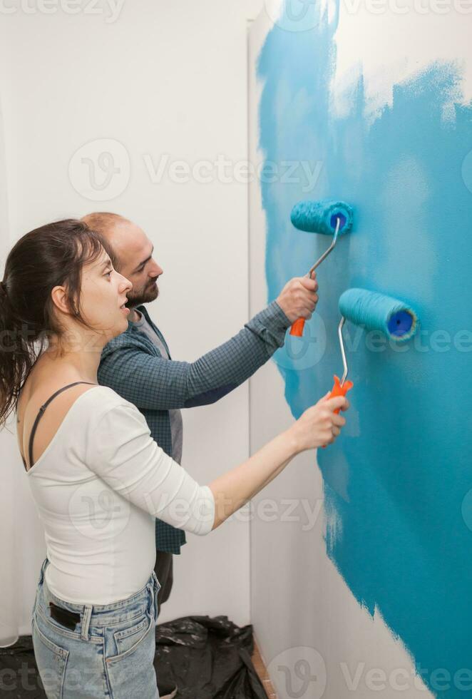 gelukkig paar geven de muur een make-over met blauw verf gebruik makend van rol borstels. huis gedurende vernieuwing, decoratie en schilderen. interieur appartement verbetering onderhoud. rol, ladder voor huis reparatie foto