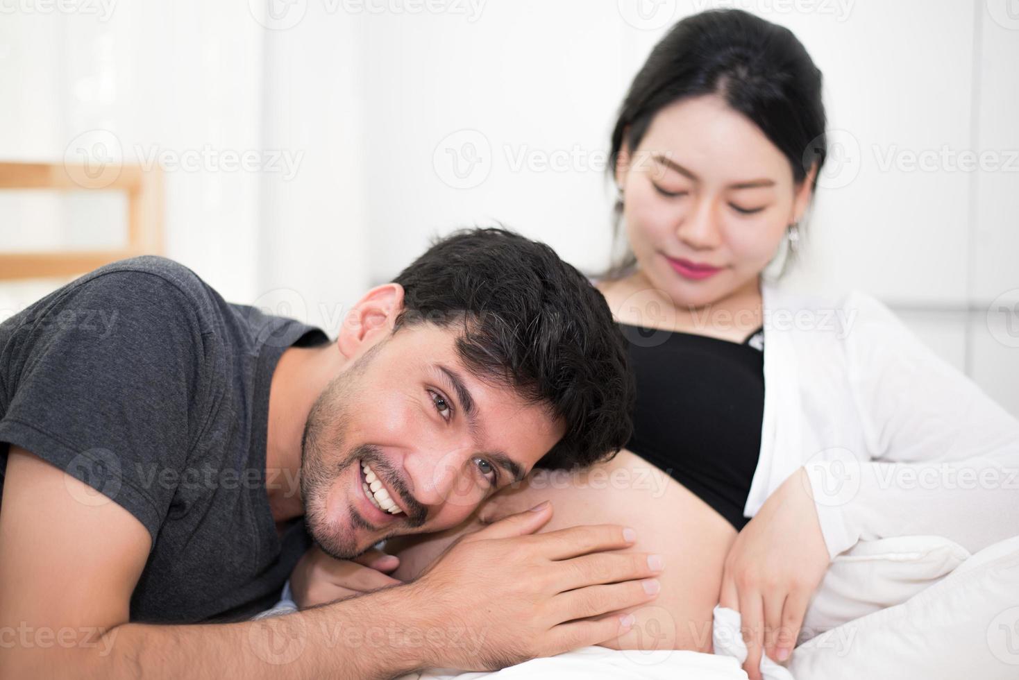 vader hoort zijn zoon of dochter schoppend geluid in de buik van de moeder wanneer hij thuis op bed ligt. familie en liefhebbers concept. gelukkig zoet huis en bruiloftsthema foto